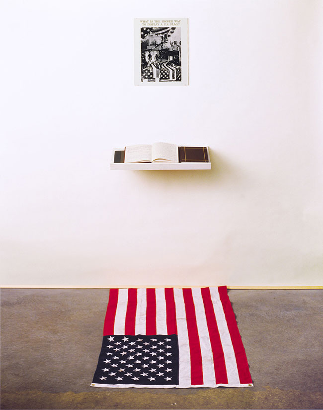 Dread Scott. "What is the Proper Way to Display a U.S. Flag?”, 1988. Installation for audience participation: Silver gelatin print, books, pens, shelf, active audience, US flag; 80 x 28 x 12 inches. Courtesy the artist.
