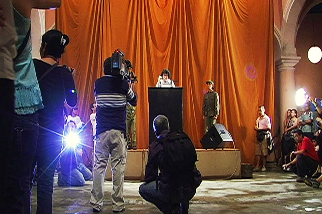 Tania Bruguera. El susurro de Tatlin #6 (Versión La Habana) / Tatlin’s Whisper #6 (Havana version), 2009. Decontextualization of an action: stage, podium, microphones, one loudspeaker inside and one loudspeaker outside of the building, two persons dressed in military outfits, white dove, one minute free of censorship per speaker, 200 disposable cameras with flash; dimensions variable. Performance view: Tenth Havana Biennial, Central Patio of the Wifredo Lam Contemporary Art Center, Havana, Cuba. Video documentation of performance: HD video; 40 minutes 30 seconds; edition of 5 (+ 1 AP). Courtesy Studio Bruguera. © Tania Bruguera