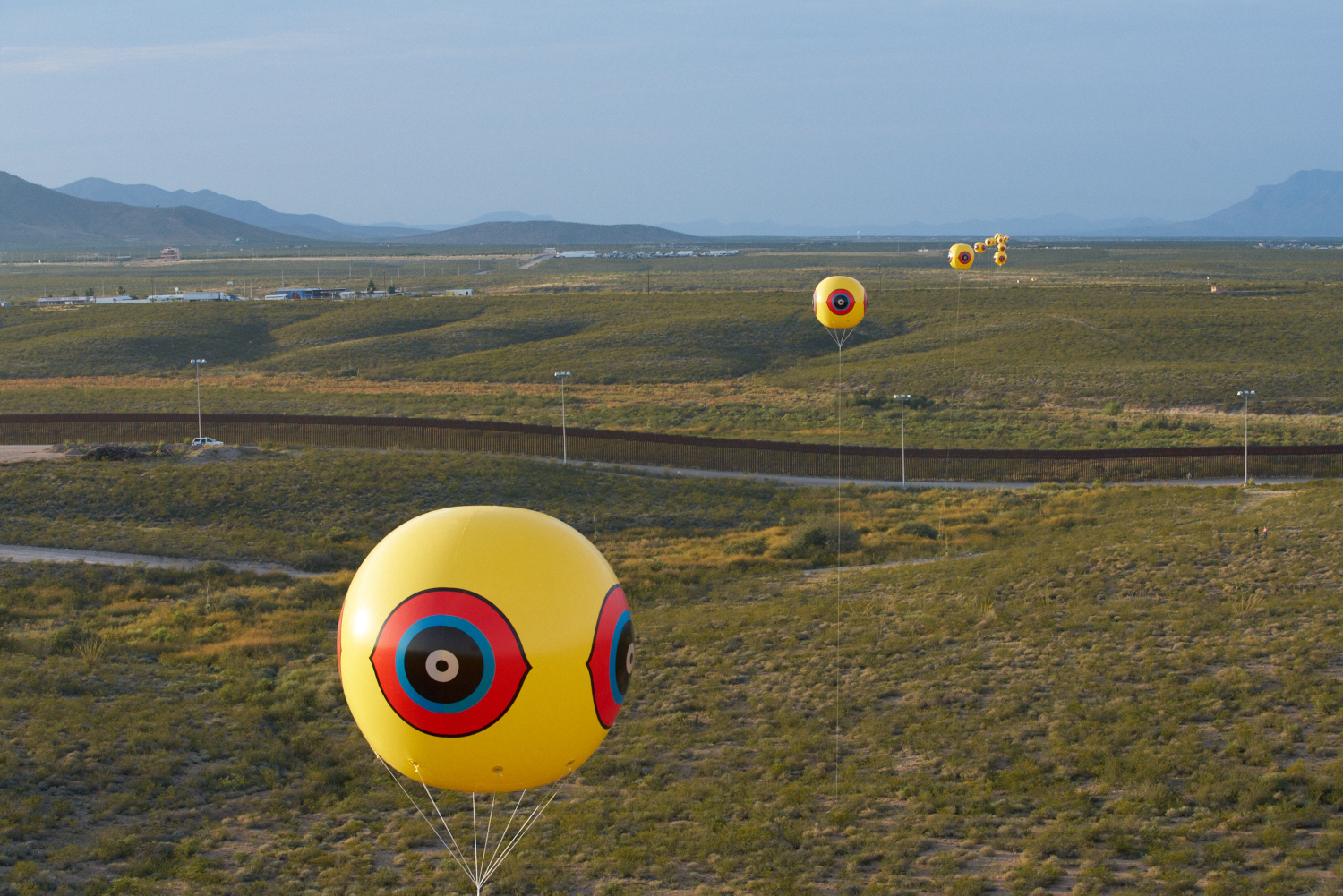 Installation detail, seen from Mexico, showing one of the balloons that comprise the Repellent Fence / Valla Repelente in relationship to the US border fence. In this image the US Border Fence reflects the sunlight at dusk. Photo by Michael Lundgren. Courtesy of Postcommodity.