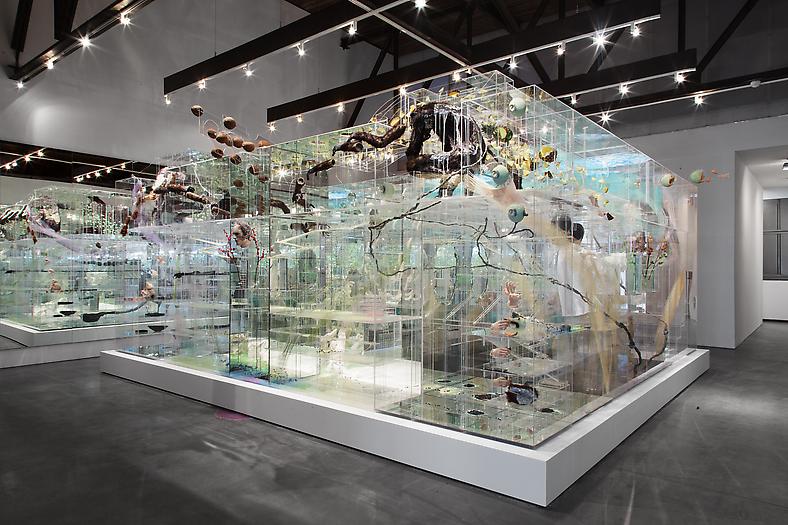 David Altmejd. "The Flux and The Puddle," 2014. Plexiglas, quartz, polystyrene, expandable foam, epoxy clay, epoxy gel, resin, synthetic hair, clothing, leather shoes, thread, mirror, plaster, acrylic paint, latex paint, metal wire, glass eyes, sequin, ceramic, synthetic flowers, synthetic branches, glue, gold, feathers, steel, coconuts, aqua resin, burlap, lighting system including fluorescent lights, Sharpie ink, wood 129 x 252 x 281 inches (327.7 x 640.1 x 713.7 cm). Courtesy of the artist and Andrea Rosen Gallery, New York, NY.